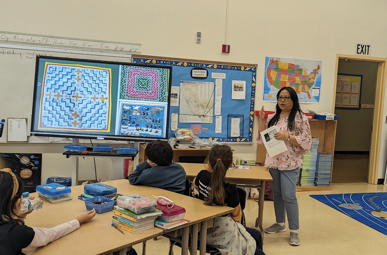 A classroom photo shows students looking ahead at a screen with colorful Hmong embroidery patterns near a whiteboard. An asian lady with long hair and glasses expressively gestures to them as she speaks and they listen