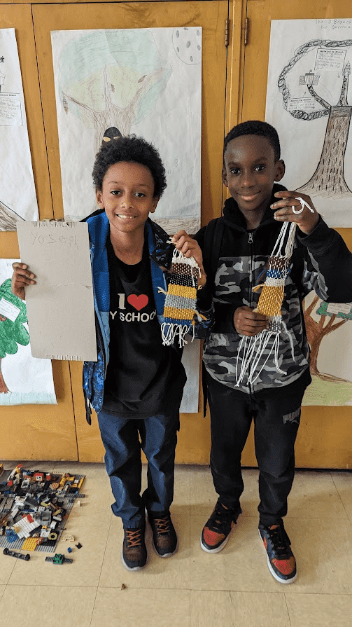Two fourth graders proudly show their multi-colored yarn-weavings. Both of them smiles at the camera, while the boy on the left holds up a cardboard loom now empty of fiber