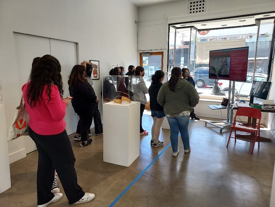 photo of 6-10 teens in an art gallery watching a screen slideshow about featured artists. There's pedestal stands for various art around them and a big window of walls looking out to the screen everyone's facing towards.