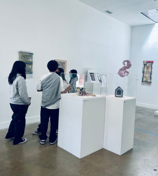 four middle schoolers in matching pants and grey hoodies chat about fiber art hung up in the craft center