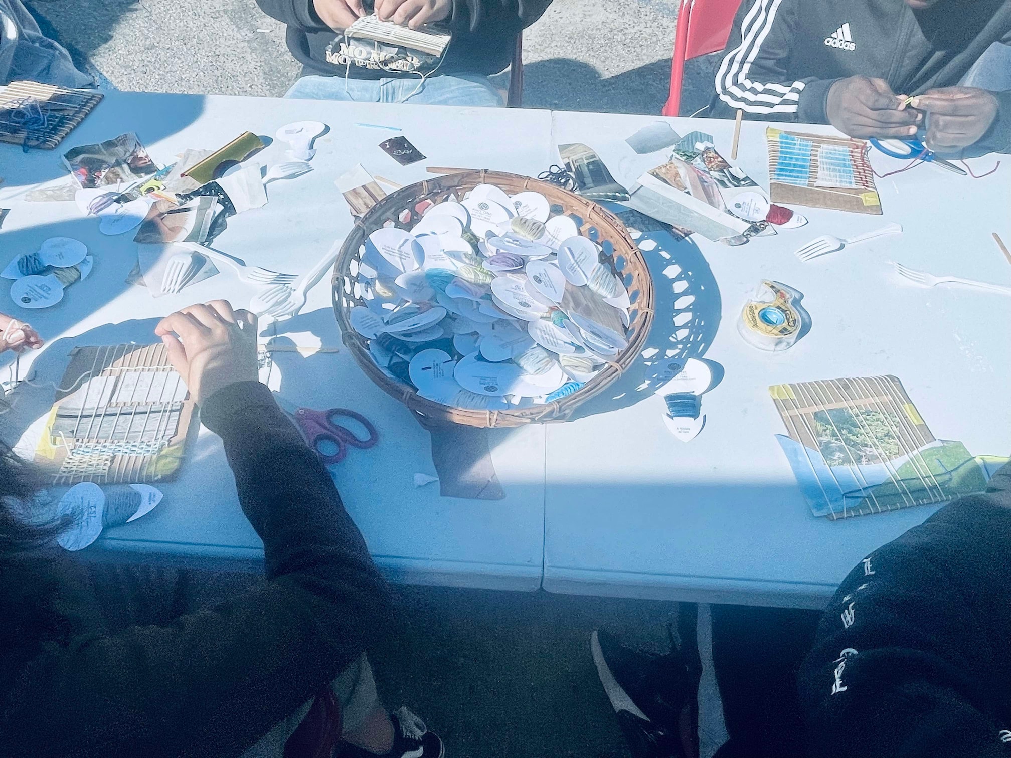 Craft table with various colorful yarn weavings, cardboard looms and a few students' arms in the frame